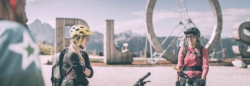 Zwei Mountainbikerinnen mit Helm vor großem ROS-Schild in den Bergen