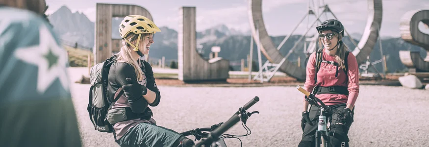 Biken á la Brixen © Brixen Tourismus - Manuel Kottersteger Zwei Mountainbikerinnen mit Helm vor großem ROS-Schild in den Bergen