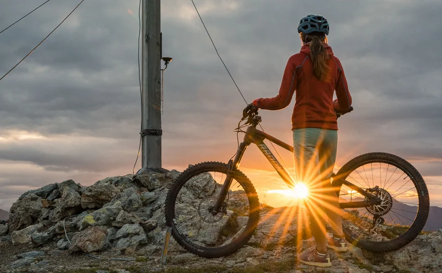 Mountainbike-Urlaub in der Region 3 Zinnen Dolomites © Elias Ochner - Stefan Schwabl Person mit Fahrrad steht neben Gipfelkreuz bei Sonnenuntergang