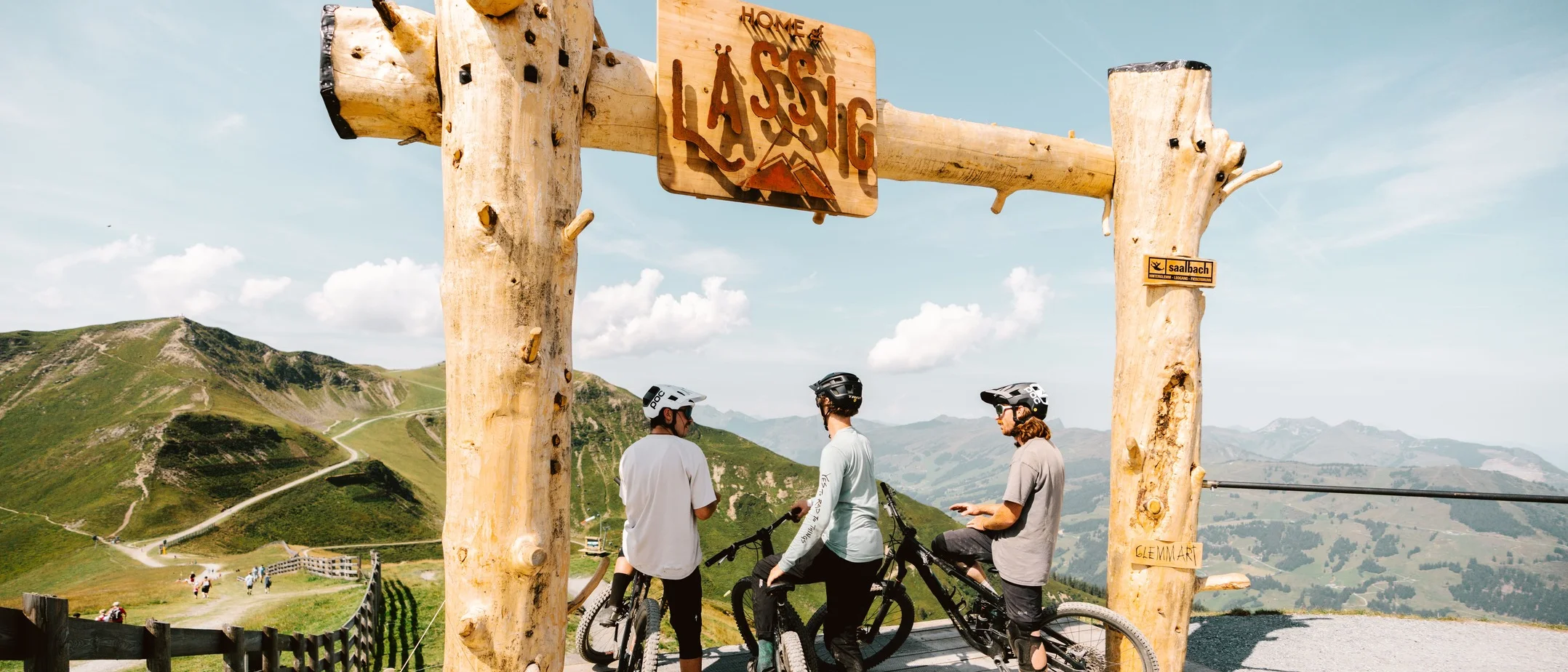 Unterwegs am Schattberg © saalbach.com/Moritz Ablinger Drei Mountainbiker unter einem Holzrahmen mit Bergsicht und blauem Himmel