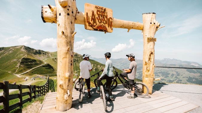 Drei Mountainbiker unter einem Holzrahmen mit Bergsicht und blauem Himmel