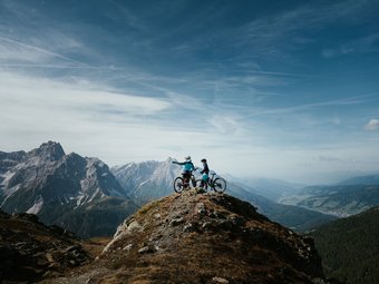 Zwei Mountainbiker auf einem Berggipfel mit Ausblick auf Gebirge