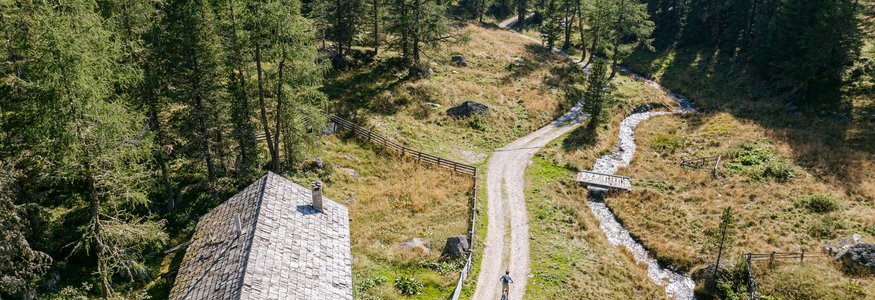 Zwei Radfahrer auf einem Weg neben einem Haus und Bach in den Bergen