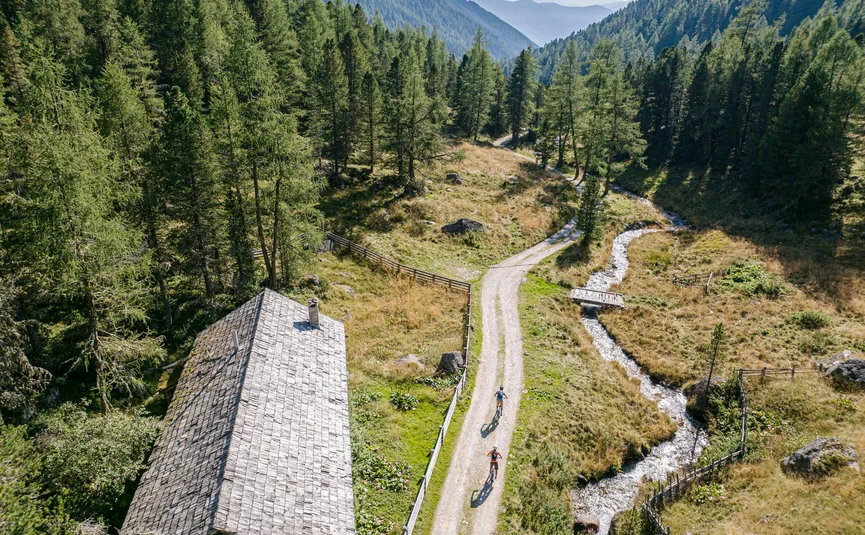 Zwei Radfahrer auf einem Weg neben einem Haus und Bach in den Bergen