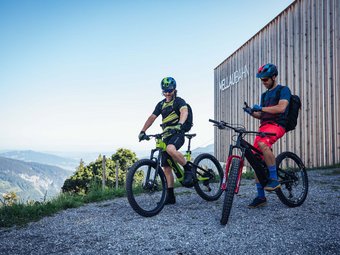 Zwei Mountainbiker mit Helmen stehen vor dem Gebäude MellauBahn in den Bergen
