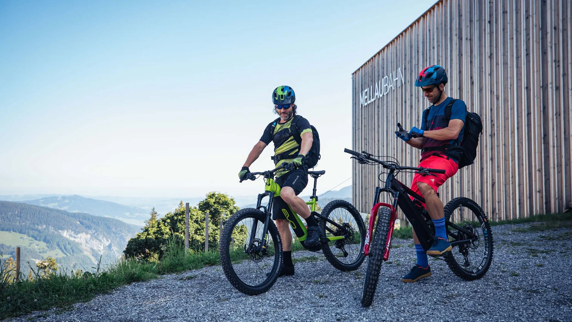 Zwei Mountainbiker mit Helmen stehen vor dem Gebäude MellauBahn in den Bergen
