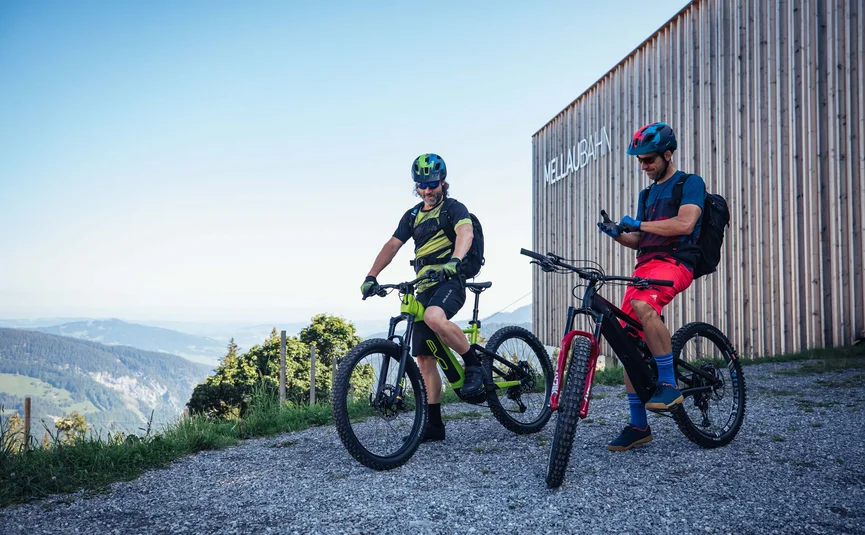 Zwei Mountainbiker mit Helmen stehen vor dem Gebäude MellauBahn in den Bergen