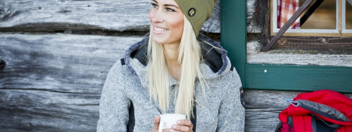 Aus 3 mach 4 - 1 Nacht geschenkt Frau in grauem Pullover und grüner Mütze sitzt mit Tasse vor Holzhütte