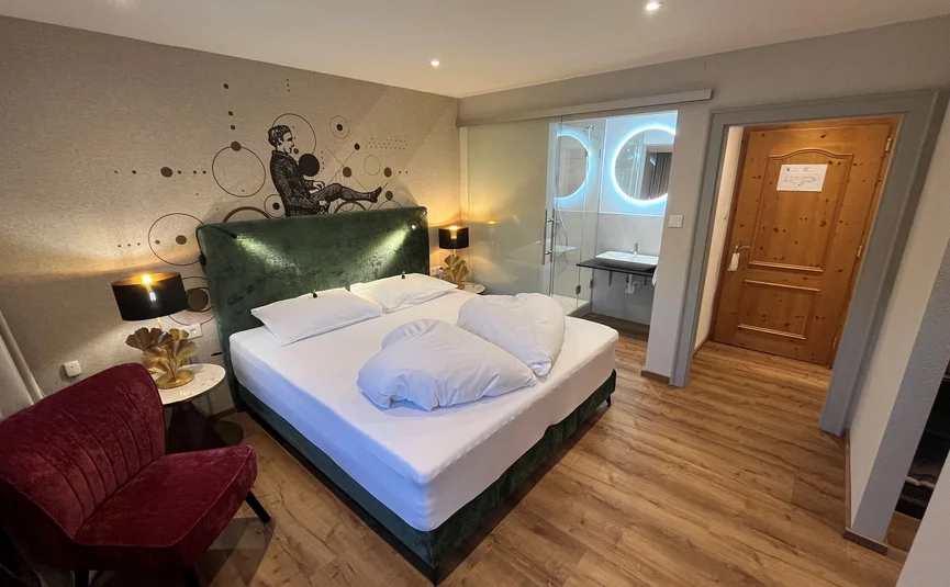 Modernes Hotelzimmer mit Doppelbett, rotem Stuhl und beleuchtetem Badezimmer