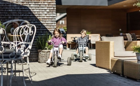 Two children riding tricycles on a patio with outdoor furniture