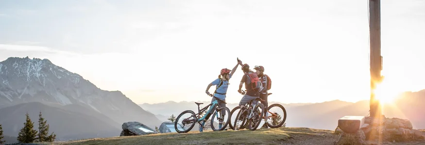 Biken & Genießen im Montanara © Salzburger Sportwelt - Dennis Stratmann Drei Mountainbiker auf Gipfel mit Holzkreuz bei Sonnenuntergang