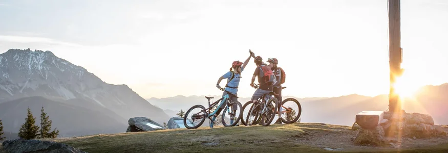 Drei Mountainbiker auf Gipfel mit Holzkreuz bei Sonnenuntergang