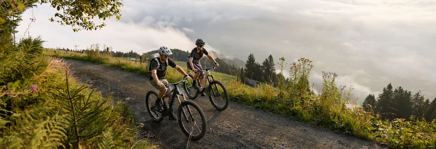 Zwei Mountainbiker auf Bergweg mit Nebel und Bergen im Hintergrund