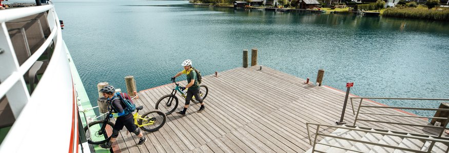 Zwei Radfahrer steigen an einem See auf eine Fähre ein, Berge im Hintergrund