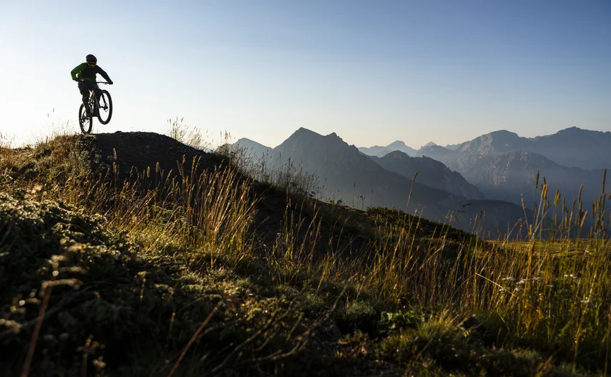 Mountainbiker springt im Sonnenlicht auf einem Bergpfad mit Alpen im Hintergrund