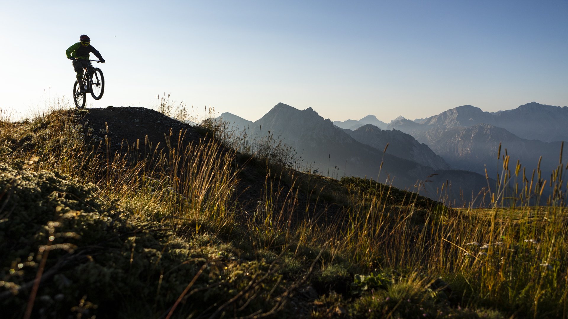 Mountainbiker springt im Sonnenlicht auf einem Bergpfad mit Alpen im Hintergrund