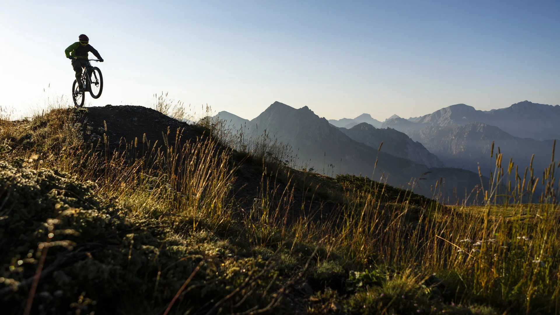 Mountainbiker springt im Sonnenlicht auf einem Bergpfad mit Alpen im Hintergrund
