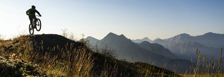 Mountainbiker springt im Sonnenlicht auf einem Bergpfad mit Alpen im Hintergrund