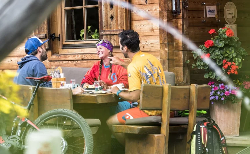 Three cyclists laughing while sitting at a wooden table outside a cabin