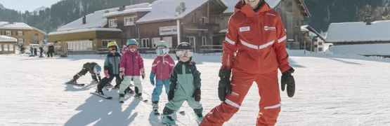 Winterurlaubswoche Skilehrer in rotem Anzug fährt mit Kindern auf einer sonnigen Piste