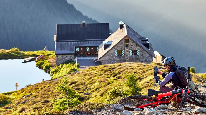 Mountainbiker macht Pause auf einem steinigen Weg mit Berghütte und See im Hintergrund