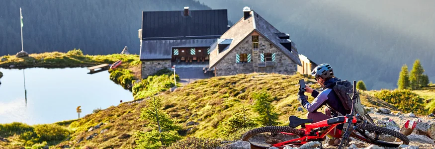 Mountainbiker macht Pause auf einem steinigen Weg mit Berghütte und See im Hintergrund