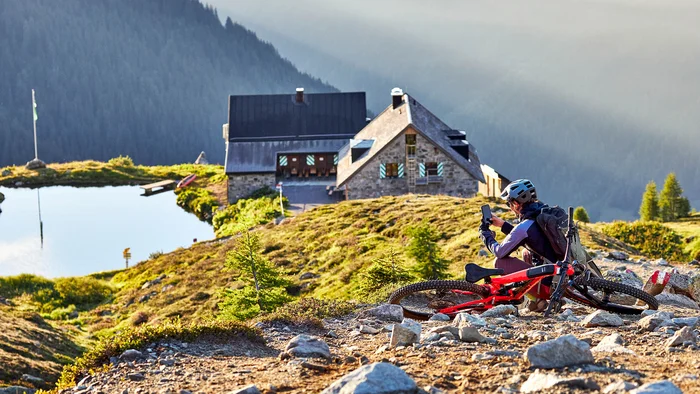Mountainbiker macht Pause auf einem steinigen Weg mit Berghütte und See im Hintergrund