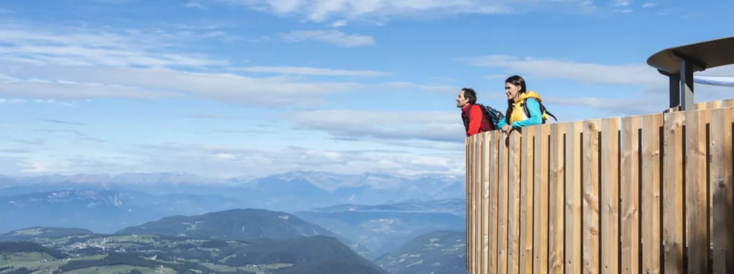 Last Minute in die Berge – 10 % Vorteil sichern! Zwei Wanderer blicken von einem hölzernen Aussichtspunkt auf ein Bergtal