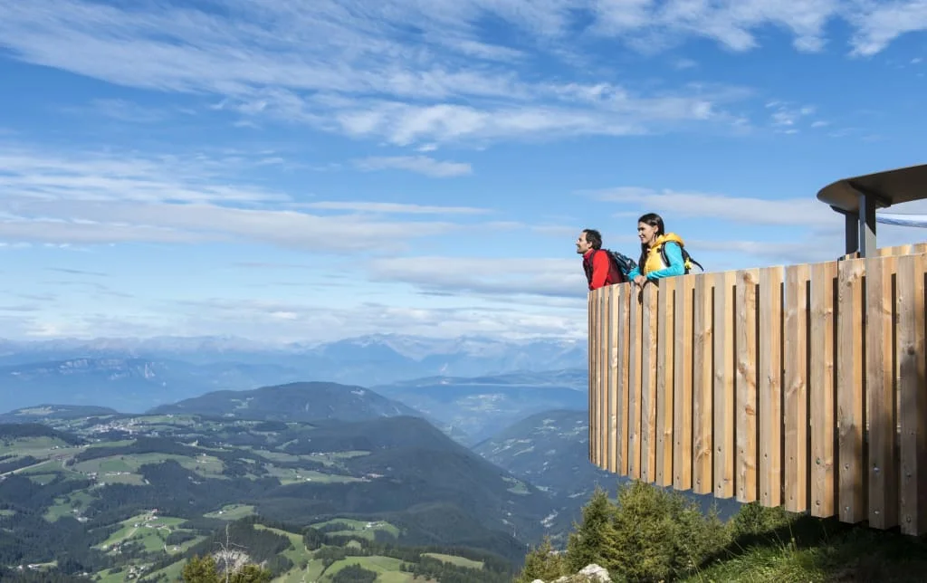 Zwei Wanderer blicken von einem hölzernen Aussichtspunkt auf ein Bergtal