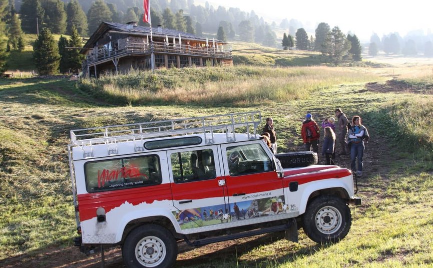 Geländewagen vor Berghütte mit Gruppe von Wanderern auf Wiese