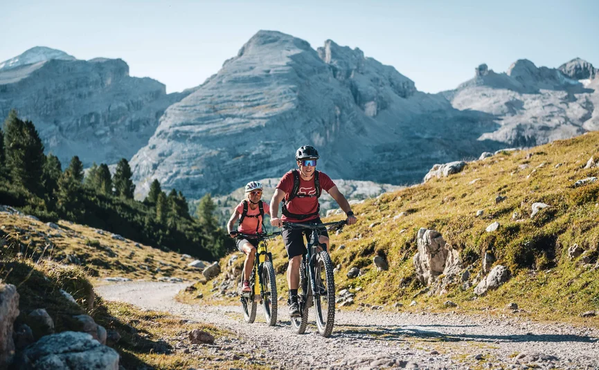 Two mountain bikers riding on a gravel trail with mountains in the background