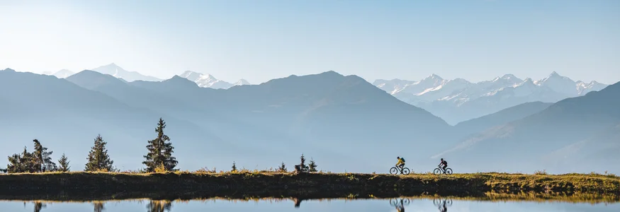 Neuer Flow im Brixental: Der Kirchberg Trail eröffnet © TVB Kitzbüheler Alpen Brixental - Mathäus Gartner Zwei Radfahrer fahren an einem Bergsee mit Bergpanorama bei klarem Himmel vorbei