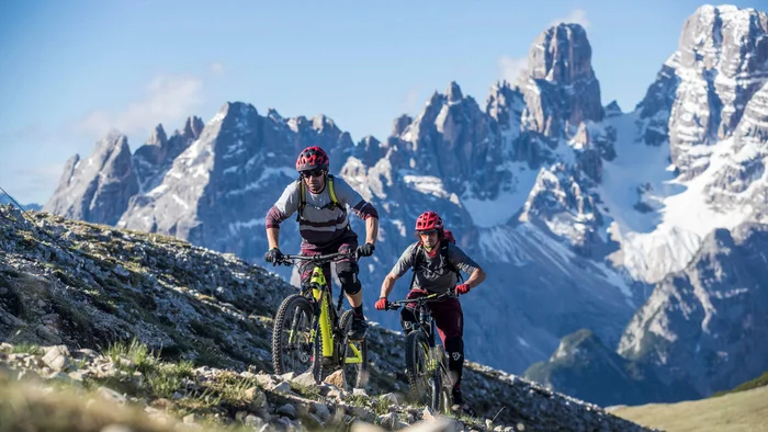 Touren & Trails © Markus Greber Zwei Mountainbiker fahren auf einem bergigen Pfad mit schneebedeckten Gipfeln im Hintergrund