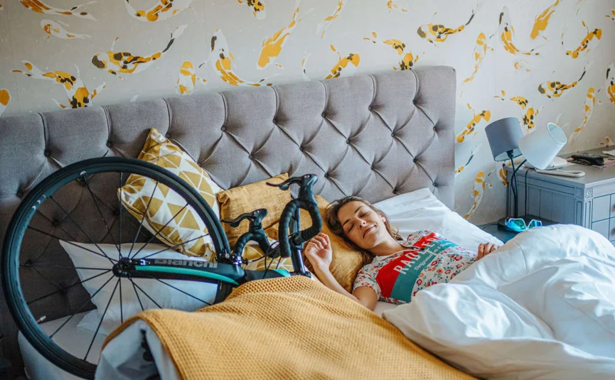 Ladylike Radmarathon Package - 2 Nächte Frau schläft im Bett neben einem Fahrrad und bunten Kissen
