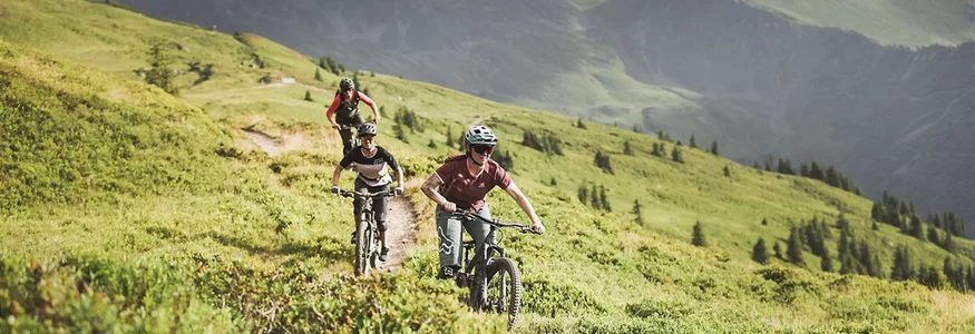 Three mountain bikers riding on a trail in green mountains under cloudy sky