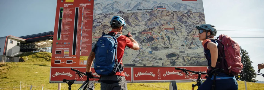 Zwei Mountainbiker sehen auf eine große KitzSki-Karte mit Routen und Lifts.