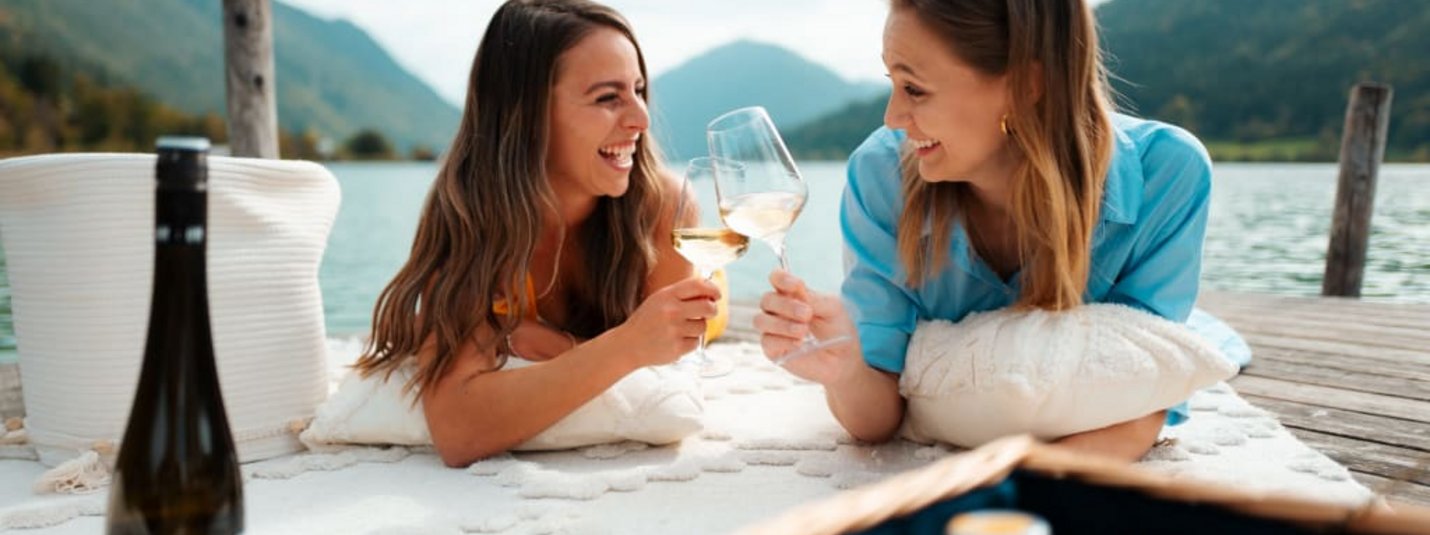 Twee vrouwen proosten met wijn aan het meer, ontspannen op een deken met picknickmand