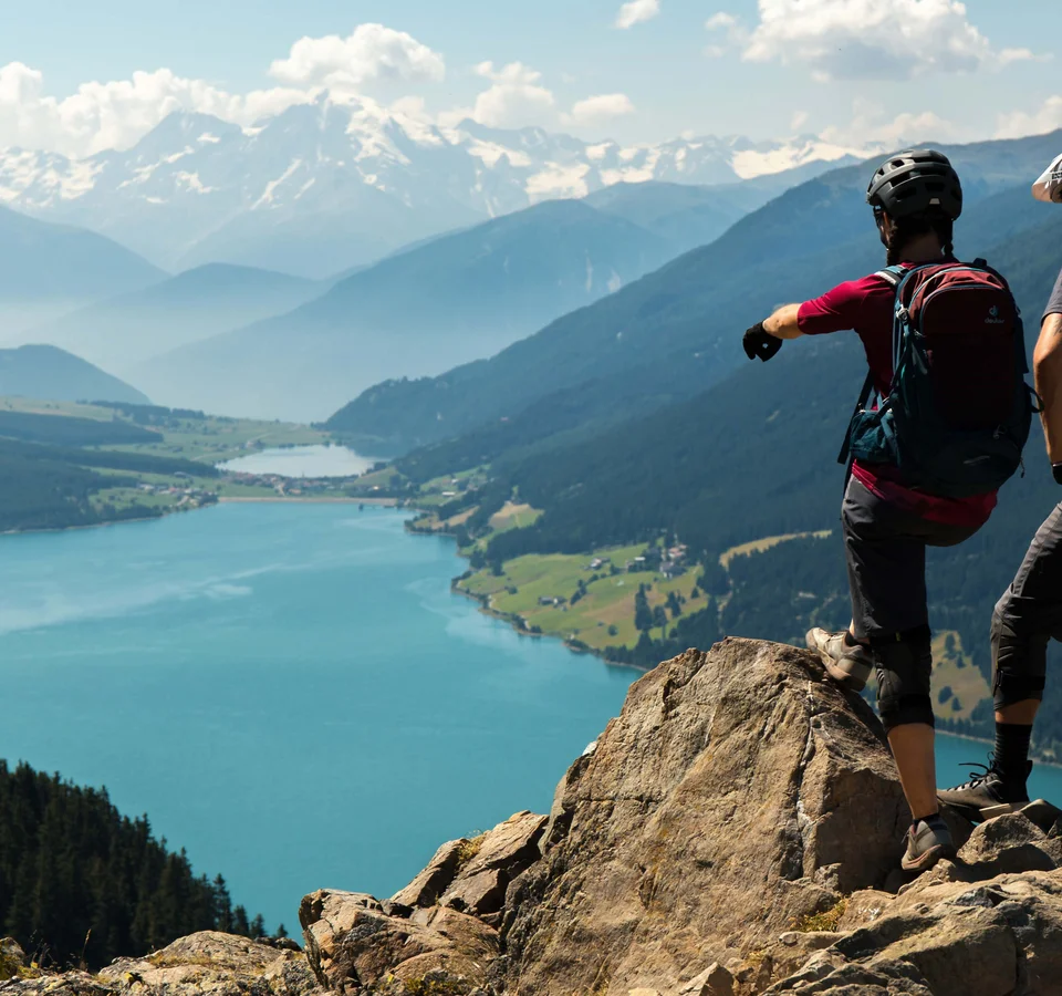 Zwei Wanderer mit Rucksäcken auf Felsen über blauem Bergsee