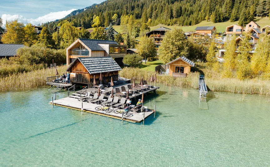 Holzsteg mit Liegestühlen am klaren See vor Berglandschaft und Häusern