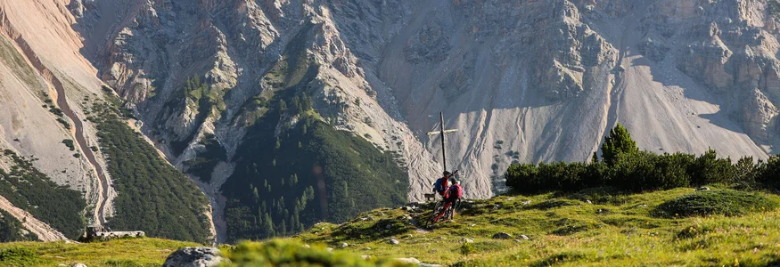 Zwei Radfahrer an einem Gipfelkreuz vor felsigen Bergen im Sonnenlicht