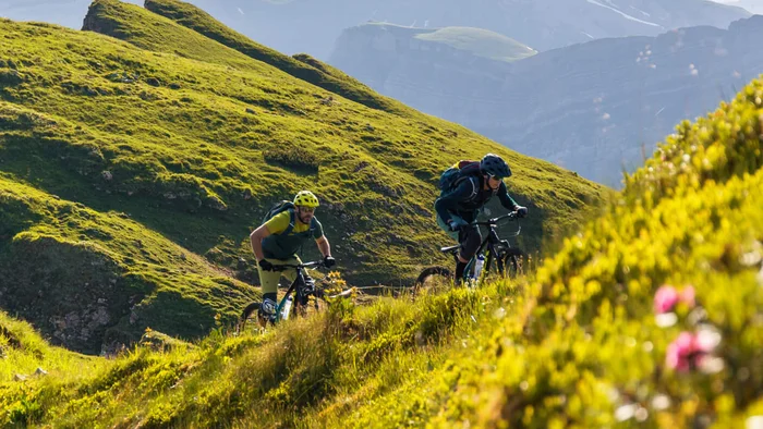 Die besten Touren & Trails der Alpen © David Karg Zwei Mountainbiker fahren auf einem grasbewachsenen Bergpfad bei sonnigem Wetter