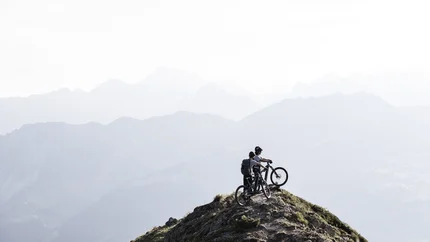 Zwei Mountainbiker auf einem Berggipfel mit nebeligen Berglandschaften im Hintergrund