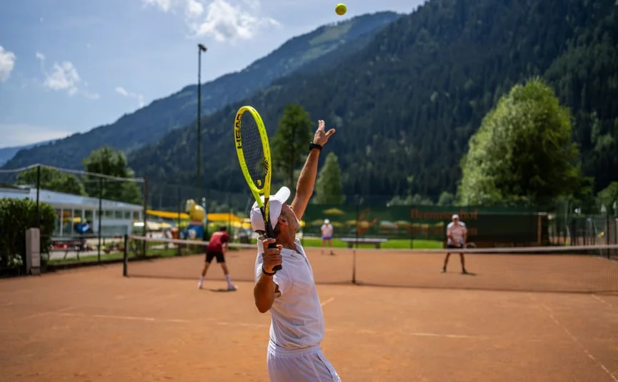 Tennisspieler serviert auf einem Außenplatz mit Bergkulisse