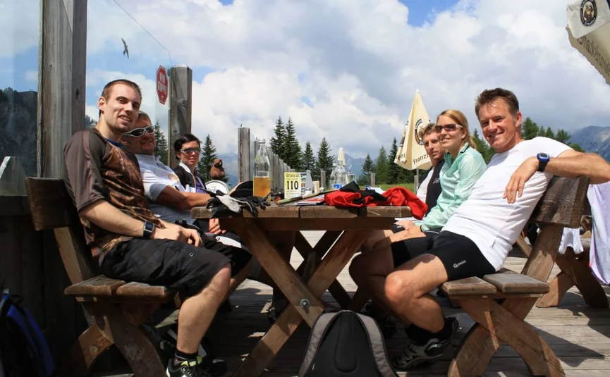 Group of six people sitting outdoors at a wooden table with mountain scenery