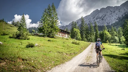 Fietser op bergpad met huis, dennenbomen en rotsachtige bergen