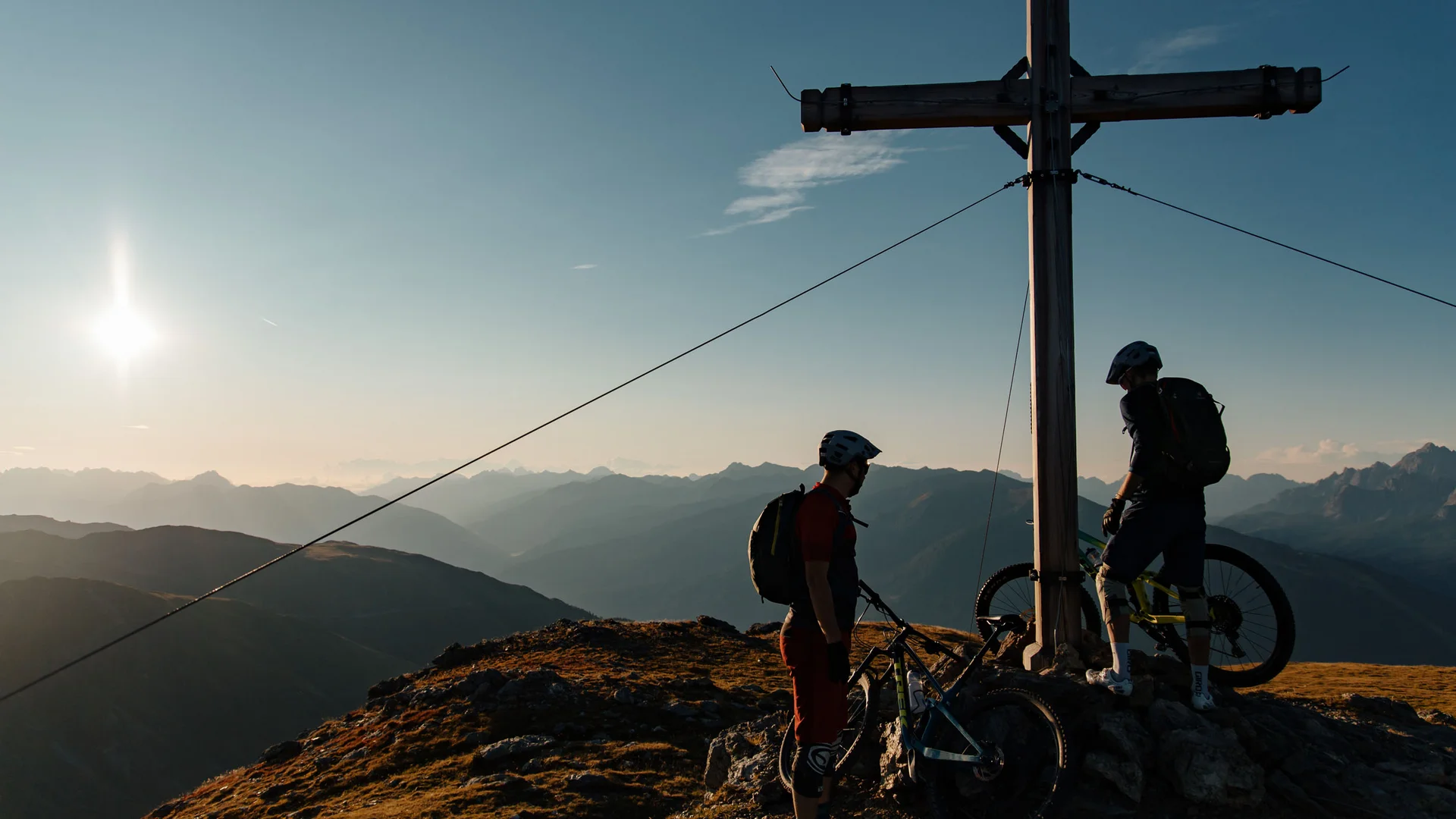 Twee mountainbikers bij een topkruis bij zonsondergang in de bergen