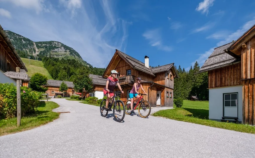 Two cyclists riding through a mountain village with wooden houses under blue sky.