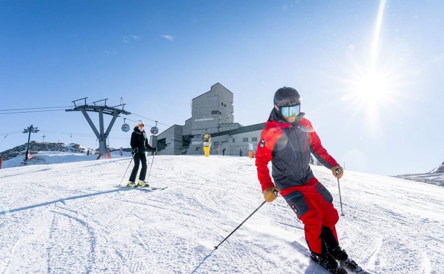 Zwei Skifahrer auf verschneitem Hang mit Seilbahn und Bergstation im Hintergrund