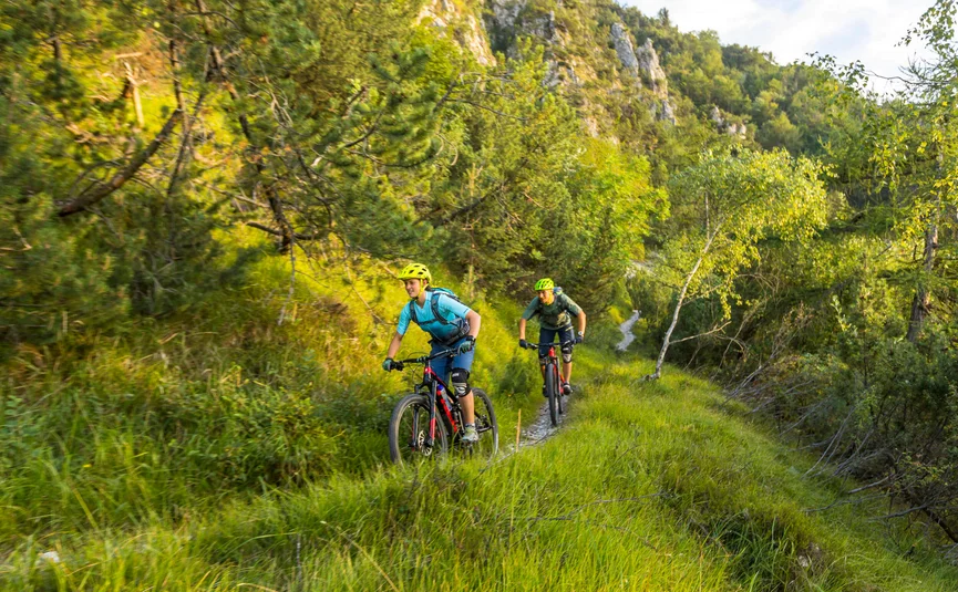 Two bikers cycling on a narrow trail in a green mountainous area