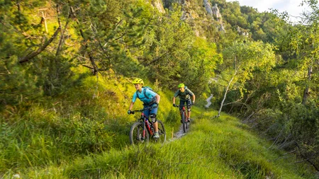 Zwei Radfahrer fahren auf einem schmalen Pfad durch eine grüne, bergige Landschaft